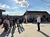 En folksamling står samlad på en landsväg mellan hus i starkt solljus.