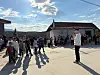 En folksamling står samlad på en landsväg mellan hus i starkt solljus.
