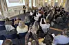 En kvinna talar från en scen inför en publik som sitter i rader av stolar och lyssnar under ett seminarium.