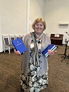 Leende kvinna håller i blå biblar med ett tryckt vitt kors i en ljus församlingssal.