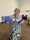 Leende kvinna håller i blå biblar med ett tryckt vitt kors i en ljus församlingssal.