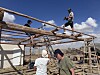 Personer monterar en takkonstruktion av trästockar utomhus under en blå himmel.