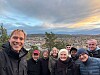 En grupp människor tar en selfie på en höjd med en stad i bakgrunden under en färgstark kvällshimmel.