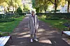 En äldre kvinna i beige trenchcoat står leende på en parkväg omgiven av träd och bänkar.