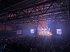 En stor publik i en mörk arena framför en färgstarkt upplyst scen med strålkastare och skärmar.