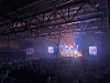En stor publik i en mörk arena framför en färgstarkt upplyst scen med strålkastare och skärmar.