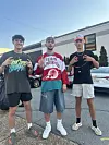 Tre unga män i streetwear och kepsar poserar på en asfalterad parkeringsplats.