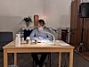 En person läser ur en stor bok vid ett bord med mikrofon i en kyrklig miljö.