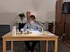 En person läser ur en stor bok vid ett bord med mikrofon i en kyrklig miljö.