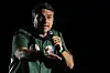 Flávio Bolsonaro talar i mikrofon och gestikulerar på en scen under en valkampanj i mörk miljö.