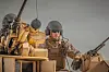 Soldat i hjälm och solglasögon på ett militärfordon utrustat med tunga vapen mot en grå himmel.