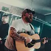 En tatuerad man i keps sjunger och spelar akustisk gitarr på en scen.