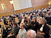 Publik som sitter på träbänkar och lyssnar under ett möte i en stor samlingssal.