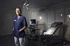 Överläkare Helena Kopp Kallner i blå arbetskläder står i ett undersökningsrum med gynekologstol på Danderyds sjukhus.