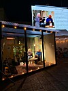Två personer i en belyst glasmonter under en utomhussändning på kvällen med en stor videoskärm i bakgrunden.