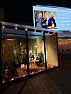 Två personer i en belyst glasmonter under en utomhussändning på kvällen med en stor videoskärm i bakgrunden.
