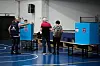 Väljare står framför blå röstbås med Israels statsvapen i en idrottshall under parlamentsvalet.
